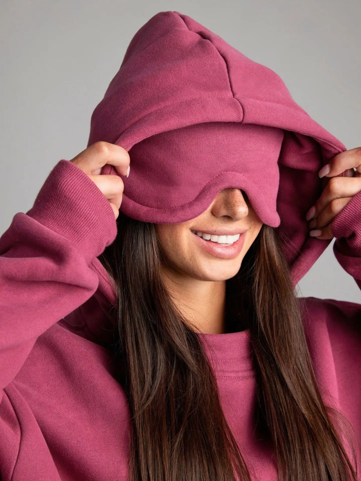 Person wearing a pink hoodie with a hood over their eyes against a gray background